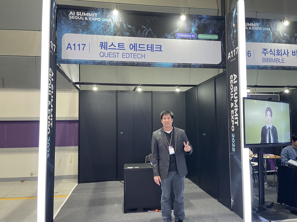 ผู้ให้คำปรึกษา AI ในงานแสดงสินค้า ชายคนหนึ่งยืนอยู่หน้าบูธ Quest Edtech ในงาน AI Summit Seoul & Expo 2025 กำลังทำท่าทางโอเค เกี่ยวกับที่ปรึกษาด้าน AI และโซลูชันการเรียนรู้ AI