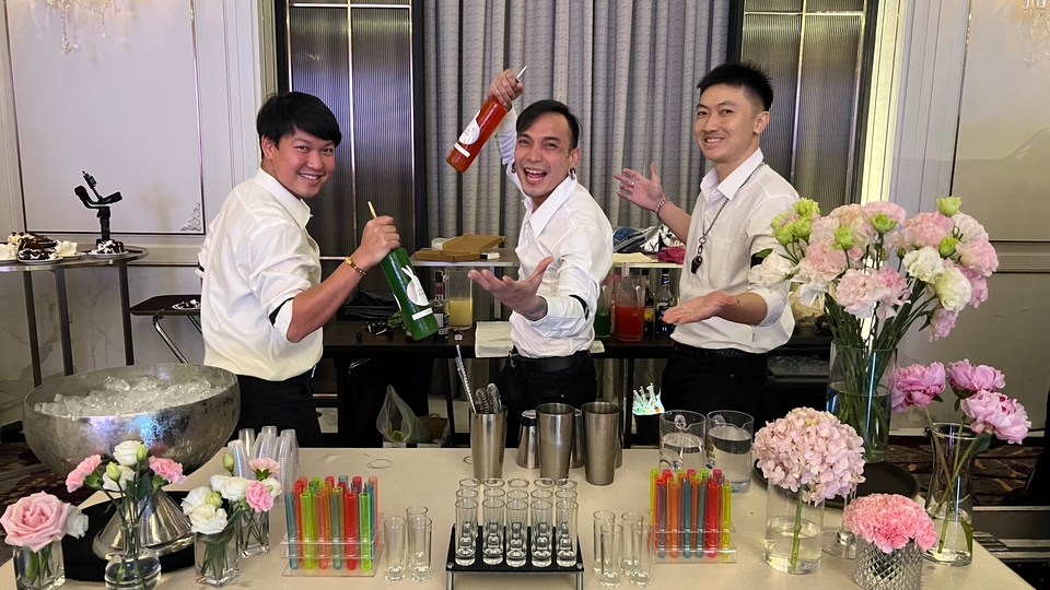 บาร์เทนเดอร์สามคนกำลังเตรียมเครื่องดื่มอย่างสนุกสนานที่บาร์ มีทั้งอุปกรณ์ผสมเครื่องดื่ม แก้ว และดอกไม้ตกแต่ง เหมาะสำหรับงานเลี้ยง อีเวนต์ หรือต้องการบาร์เทนเดอร์ส่วนตัว ติดต่อเราได้เลยเพื่อจ้างบาร์เทนเดอร์มืออาชีพ